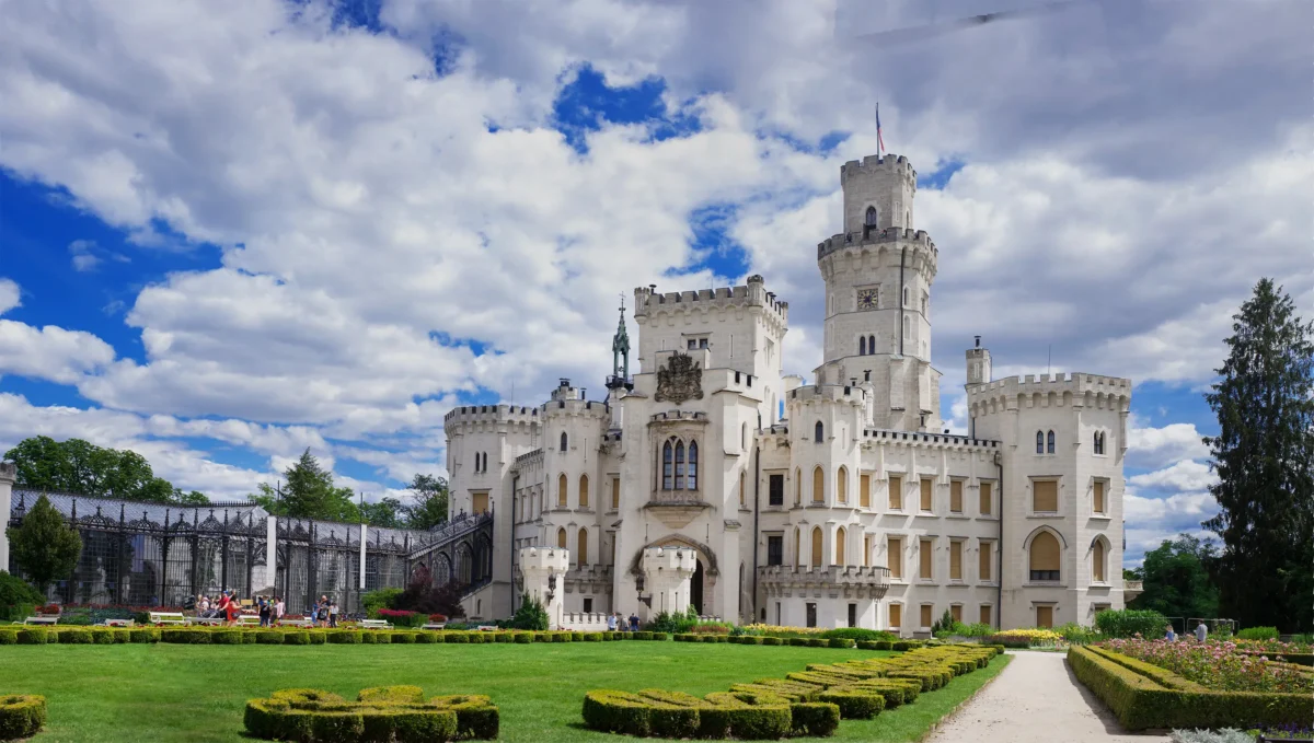 Bílý středověký hrad s cimbuřím, vlajkou na vrcholu centrální věže, zasazený do modré oblohy s mraky a formálními zahradami v přední části.