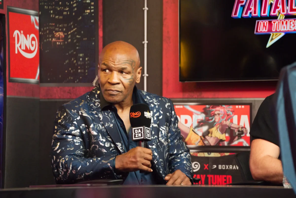 shutterstock_2622489515-změněno-na-3000x2002 1 | TVGURU.cz Mike Tyson s tetováním na obličeji v modré vzorované košili sedí u stolu a mluví do mikrofonu DAZN během televizního rozhovoru, v pozadí jsou barevné plakáty.