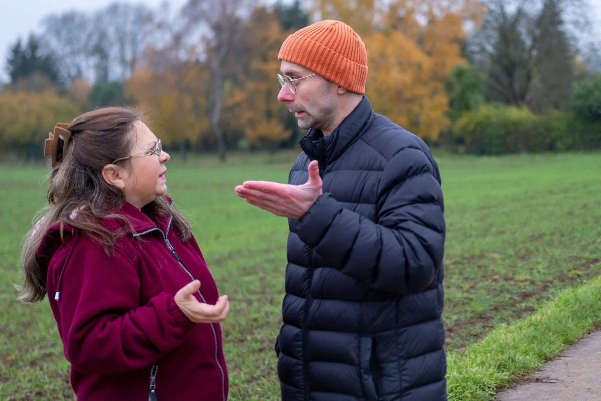 shutterstock_2650138057 | TVGURU.cz Dva lidé stojí a povídají si v travnatém parku s podzimními stromy; jeden má na sobě oranžovou pletenou čepici a černou prošívanou bundu a gestikuluje rukama na druhého, který má na sobě vínovou bundu.