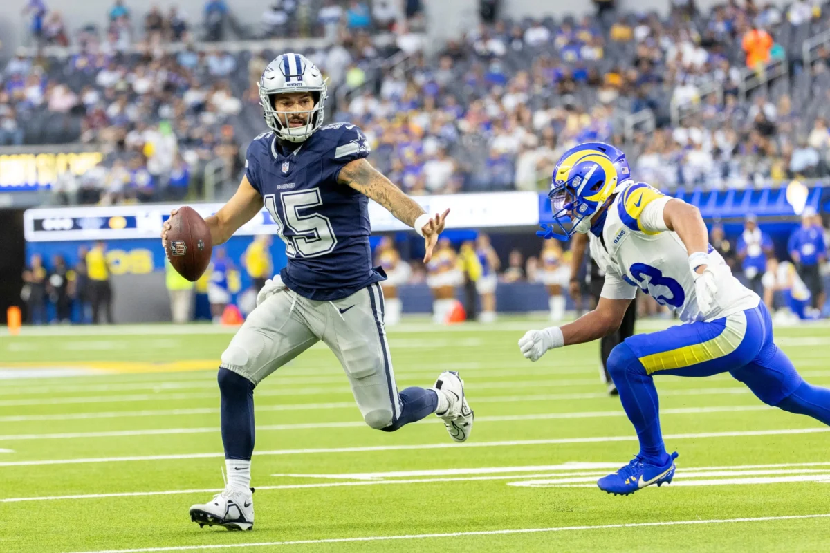 shutterstock_2664070787-změněno-na-3000x2000 | TVGURU.cz Quarterback Cowboys #15 běží s fotbalovým míčem, zatímco se obránce Rams blíží na pozadí přeplněného stadionu.