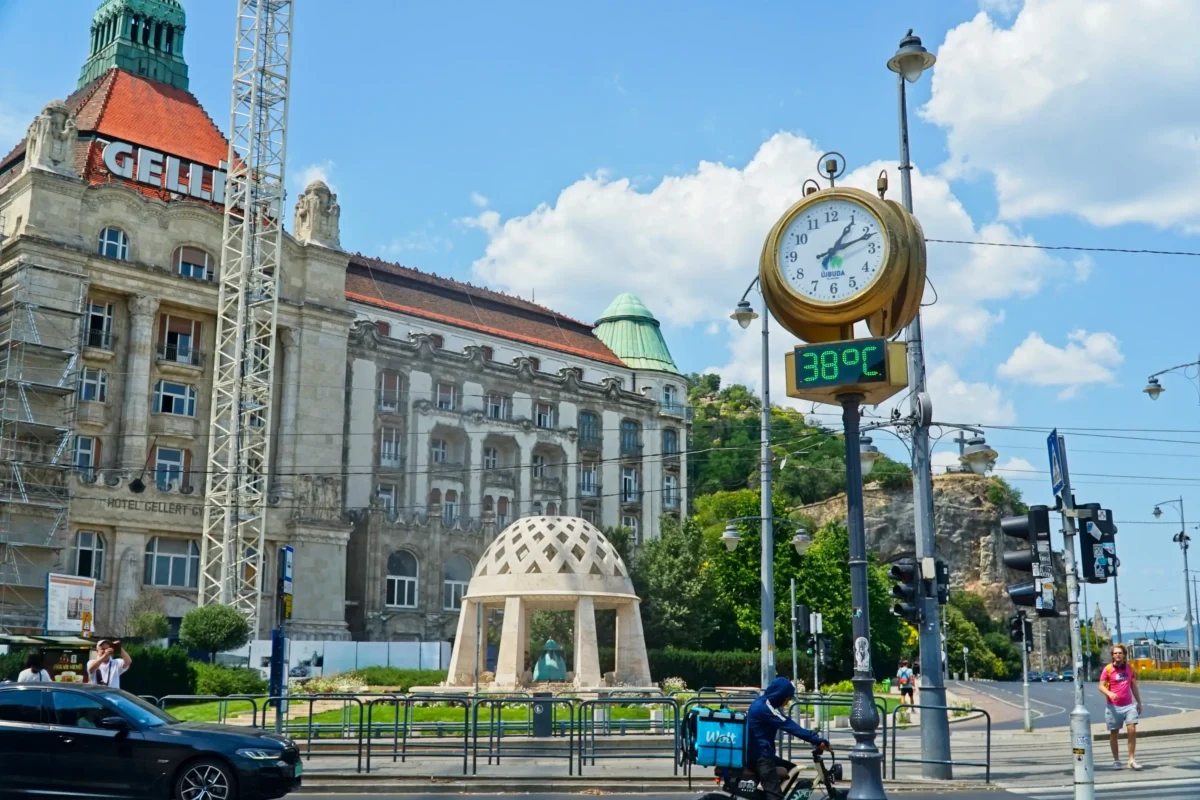Historické náměstí se starou zdobenou budovou, zelenou hodinovou věží a digitálním displejem s teplotou 38 °C pod modrou oblohou.