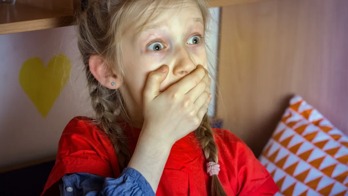 A young girl with braided hair and a red shirt covering her mouth with her hands, her eyes wide in surprise.
