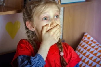 A young girl with braided hair and a red shirt covering her mouth with her hands, her eyes wide in surprise.