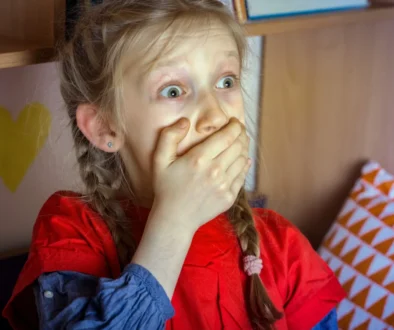 A young girl with braided hair and a red shirt covering her mouth with her hands, her eyes wide in surprise.