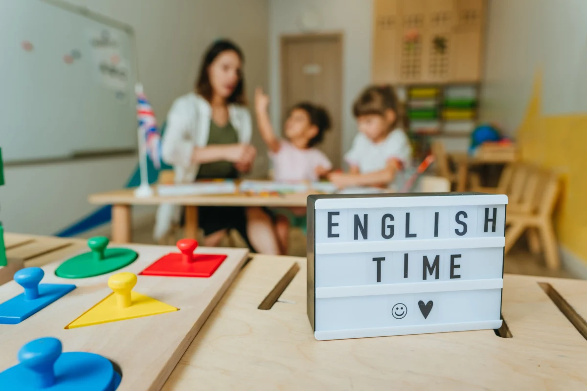 shutterstock_2186725581 1 | TVGURU.cz Teacher guiding two children at a classroom table for an English lesson; foreground shows a lightbox reading'ENGLISH TIME'.