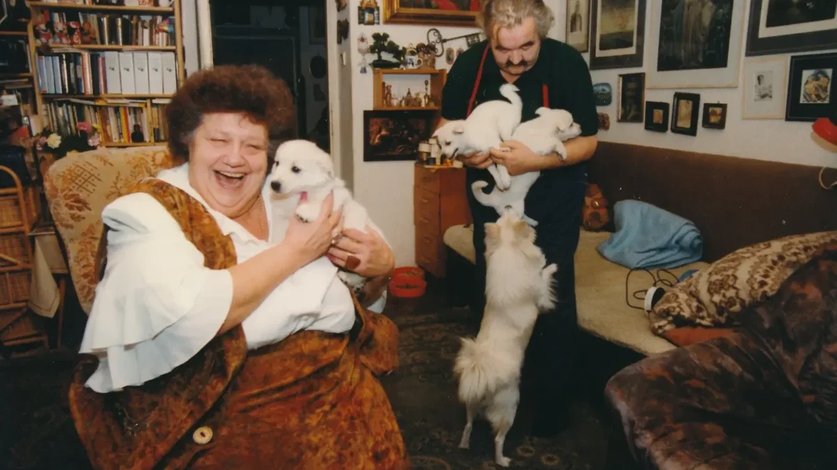 Usměvavá starší žena držící uvnitř bílé štěně, s mužem a dalšími štěňaty poblíž v útulném obývacím pokoji. - Helena Růžičková