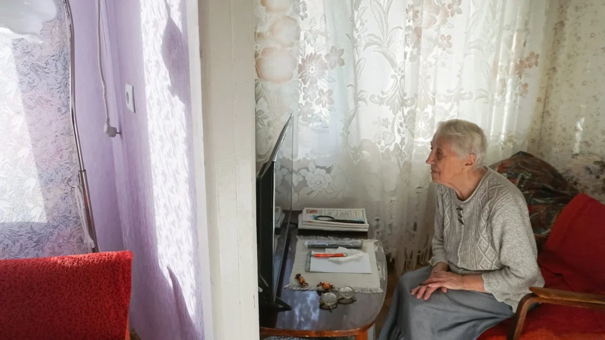 Elderly woman seated in a living room, facing a small TV on a side table with papers nearby and red upholstered chairs nearby.