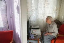 Elderly woman seated in a living room, facing a small TV on a side table with papers nearby and red upholstered chairs nearby.