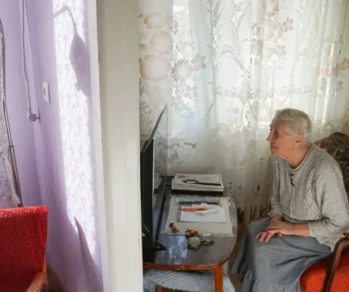 Elderly woman seated in a living room, facing a small TV on a side table with papers nearby and red upholstered chairs nearby.
