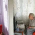 Elderly woman seated in a living room, facing a small TV on a side table with papers nearby and red upholstered chairs nearby.