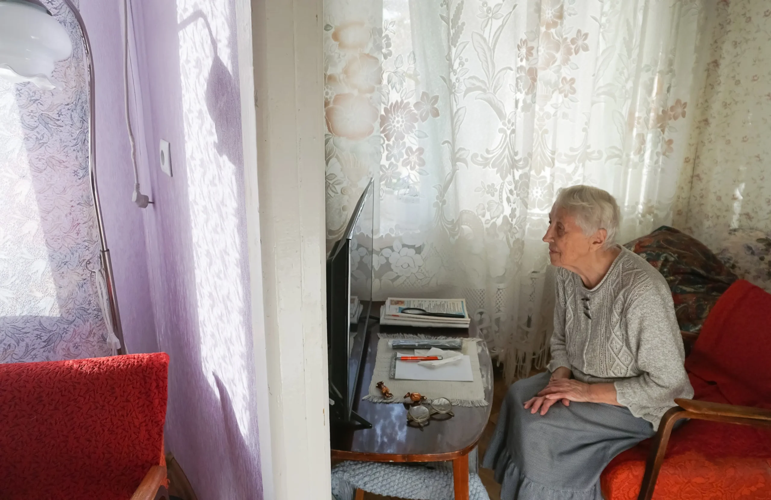 Elderly woman seated in a living room, facing a small TV on a side table with papers nearby and red upholstered chairs nearby.