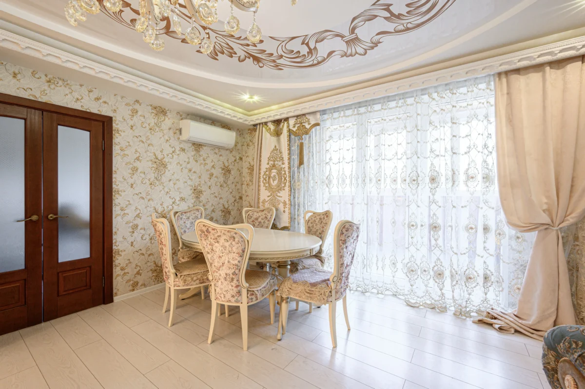 Elegant dining room with a round table and cream floral chairs, ornate wallpaper, and sheer lace curtains.