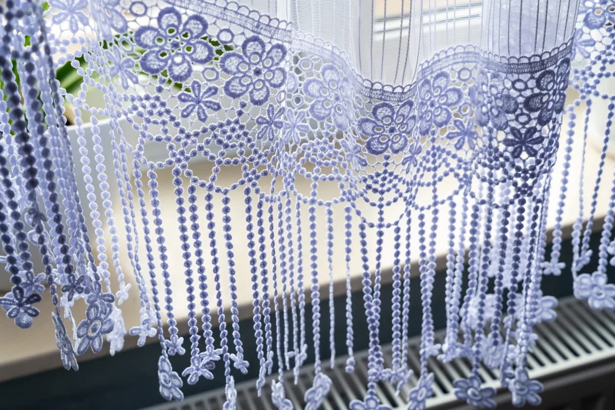 Close-up of pale blue lace curtains with floral bead patterns hanging in front of a window.