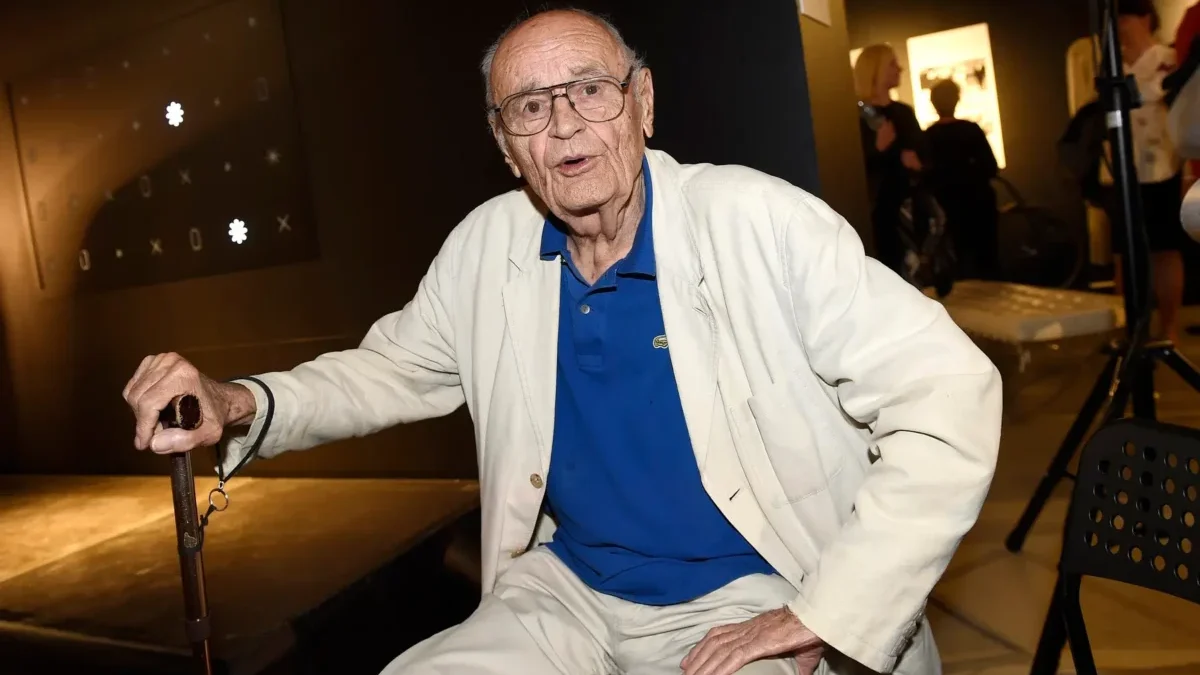 Elderly man in a white suit and blue polo sits with a cane at an indoor event.