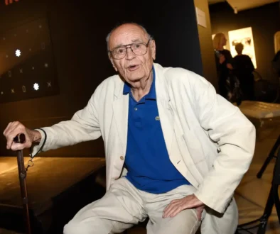 Elderly man in a white suit and blue polo sits with a cane at an indoor event.