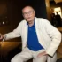 Elderly man in a white suit and blue polo sits with a cane at an indoor event.