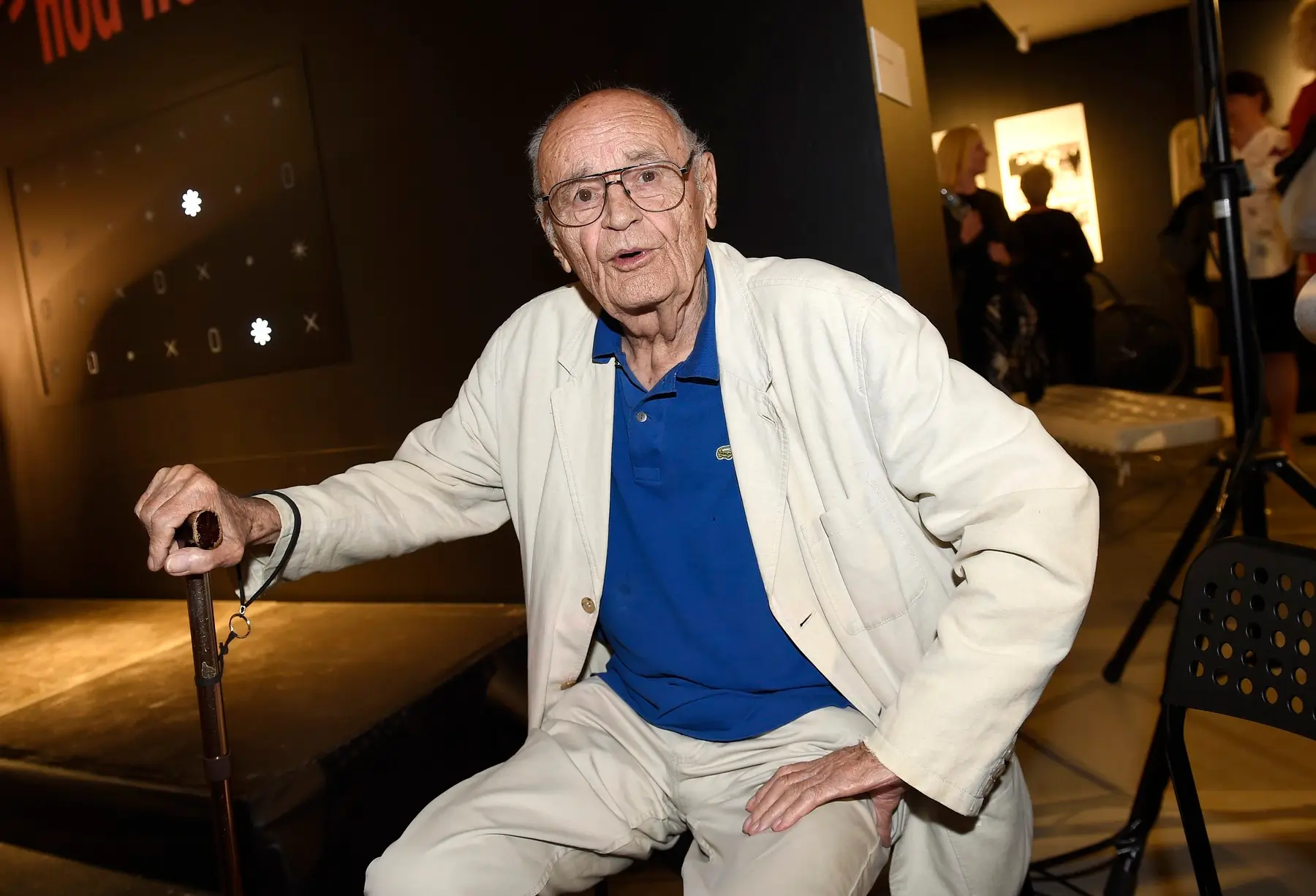 Elderly man in a white suit and blue polo sits with a cane at an indoor event.