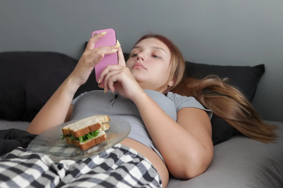 shutterstock_2760313779-2 | TVGURU.cz Slečna leží na posteli, na břiše má talíř a je na mobilu.
