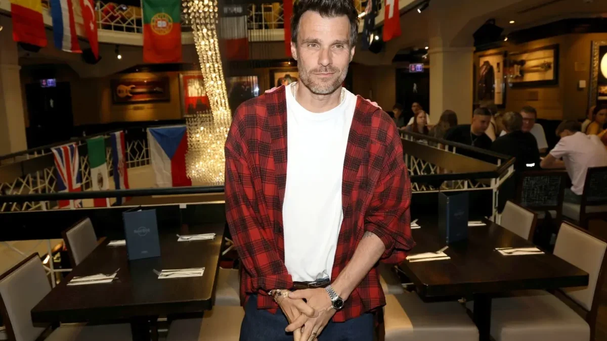 Man wearing a red plaid shirt over a white tee stands in a bustling restaurant with flags and string lights behind him.