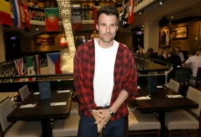 Man wearing a red plaid shirt over a white tee stands in a bustling restaurant with flags and string lights behind him.