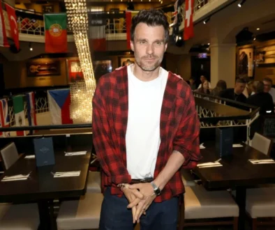 Man wearing a red plaid shirt over a white tee stands in a bustling restaurant with flags and string lights behind him.