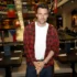 Man wearing a red plaid shirt over a white tee stands in a bustling restaurant with flags and string lights behind him.