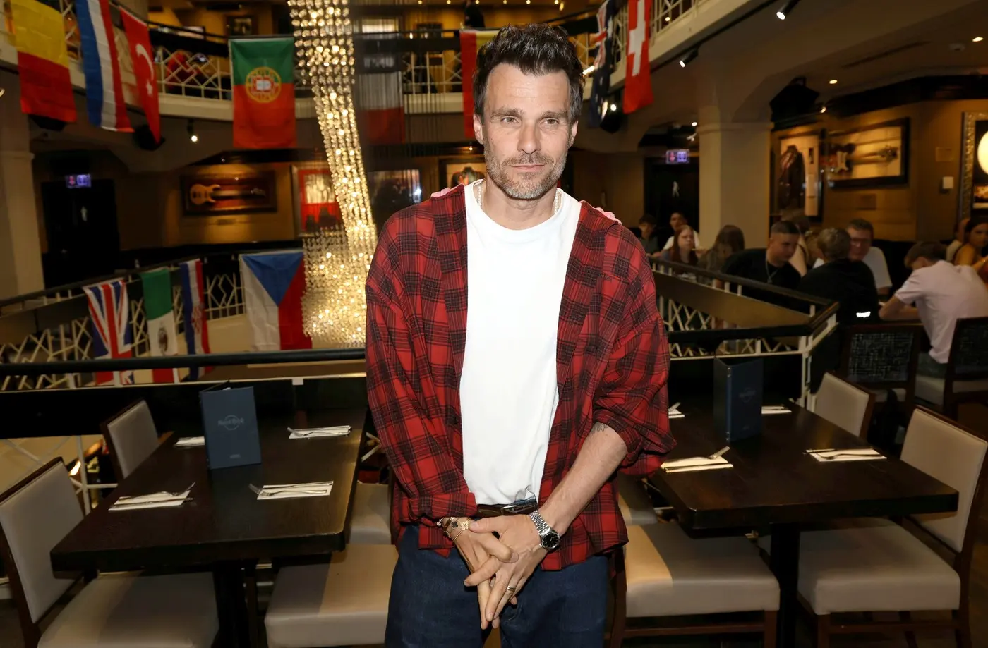 Man wearing a red plaid shirt over a white tee stands in a bustling restaurant with flags and string lights behind him.