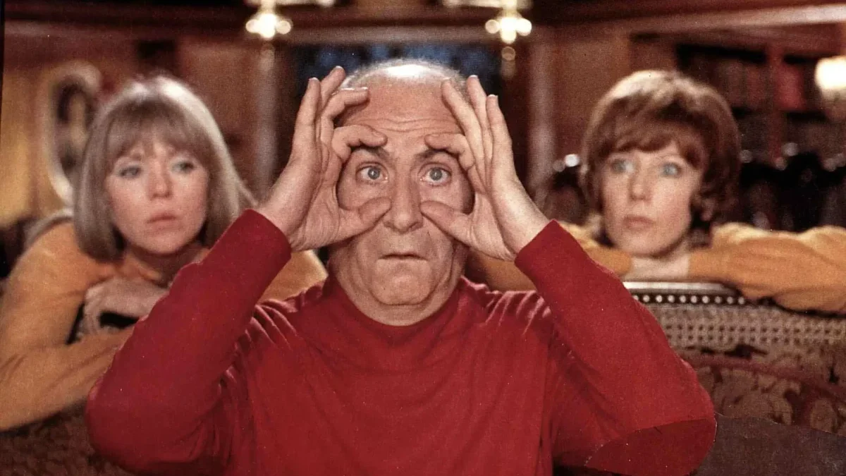 Older man in a red shirt holds his hands to his eyes like binoculars, sitting in a room with two women in the background.