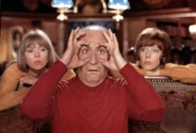 Older man in a red shirt holds his hands to his eyes like binoculars, sitting in a room with two women in the background.