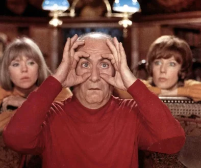 Older man in a red shirt holds his hands to his eyes like binoculars, sitting in a room with two women in the background.