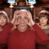 Older man in a red shirt holds his hands to his eyes like binoculars, sitting in a room with two women in the background.