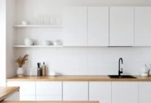 Minimalist white kitchen with wooden countertop, built-in cabinets, and open shelves holding white dishes.