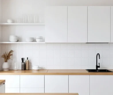 Minimalist white kitchen with wooden countertop, built-in cabinets, and open shelves holding white dishes.