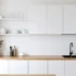 Minimalist white kitchen with wooden countertop, built-in cabinets, and open shelves holding white dishes.