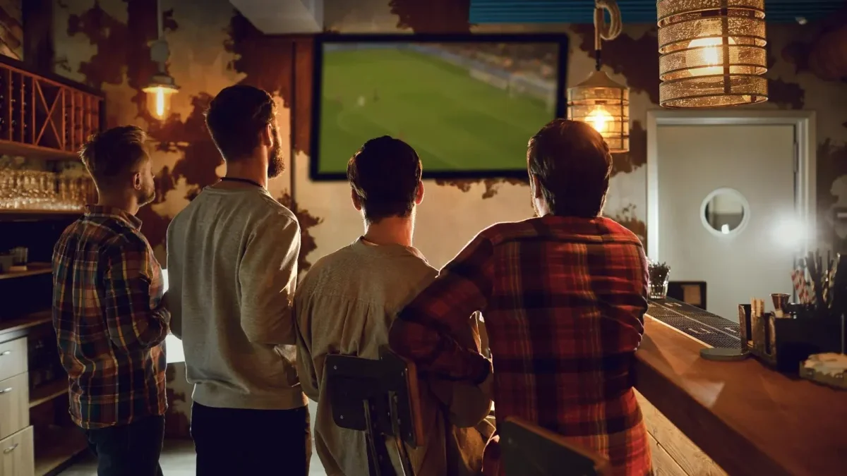 Group of men standing at a bar watching a large TV screen showing a soccer game in a dimly lit pub.