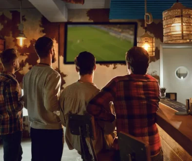 Group of men standing at a bar watching a large TV screen showing a soccer game in a dimly lit pub.
