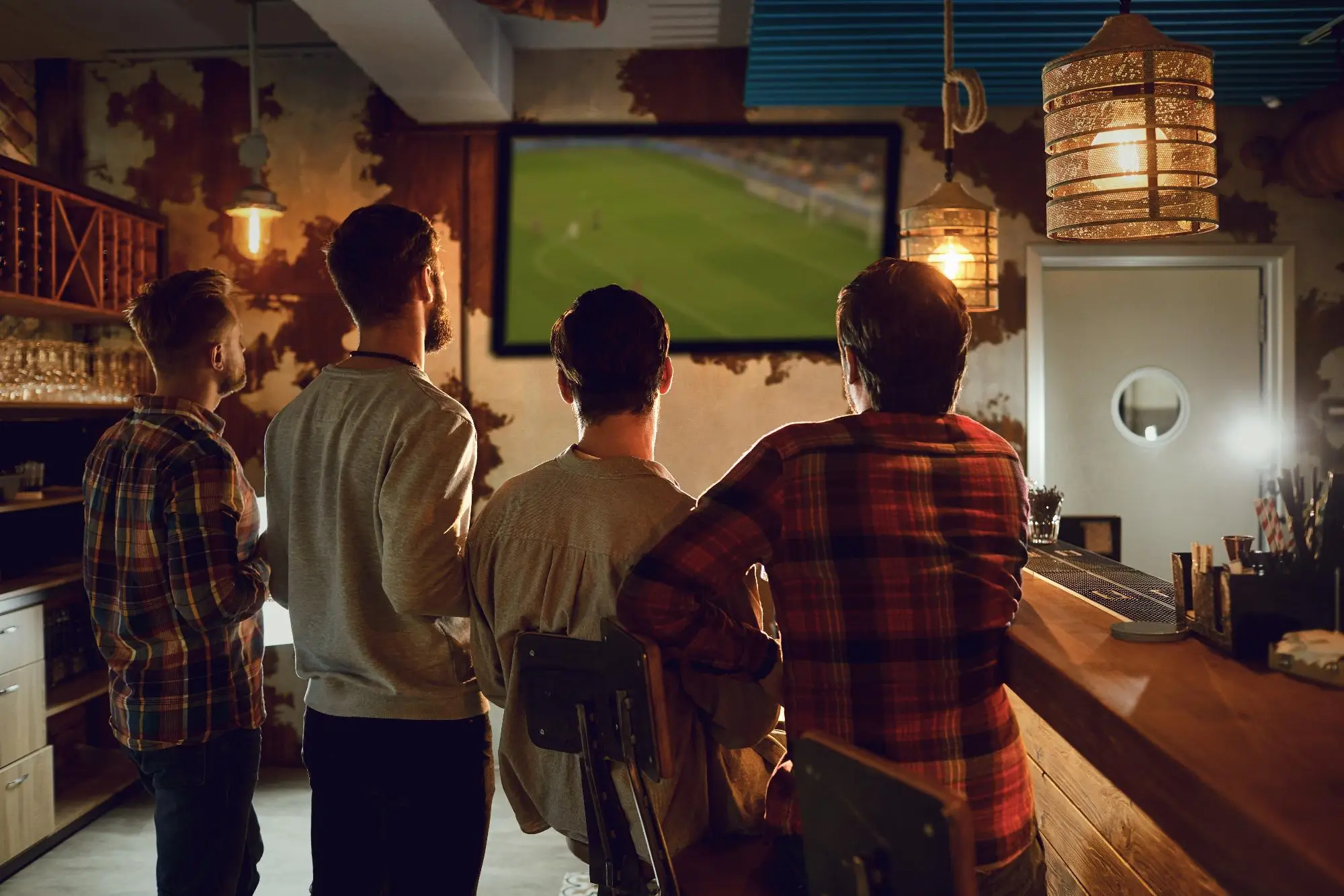 Group of men standing at a bar watching a large TV screen showing a soccer game in a dimly lit pub.