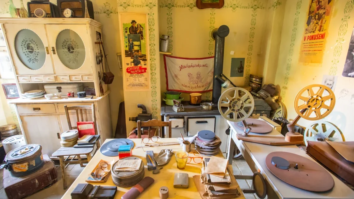 Cluttered vintage workshop with film reels, projectors, and wooden cabinets against yellow walls with retro posters and green trim patterns.