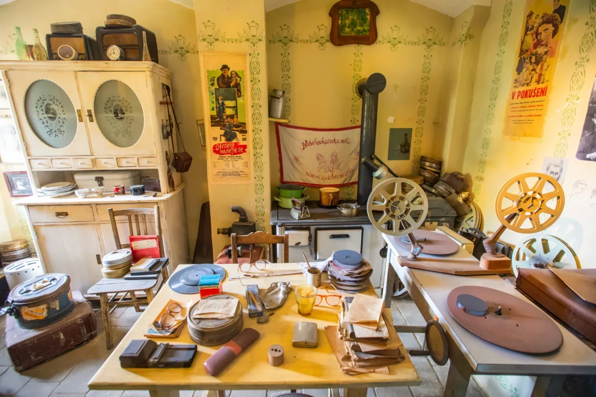 Cluttered vintage workshop with film reels, projectors, and wooden cabinets against yellow walls with retro posters and green trim patterns.