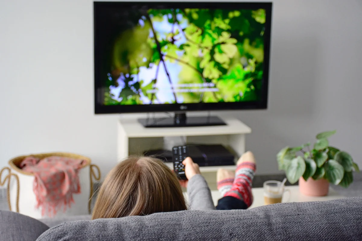 Back view of children lounging on a gray sofa, feet up, watching a wall-mounted TV in a bright living room.