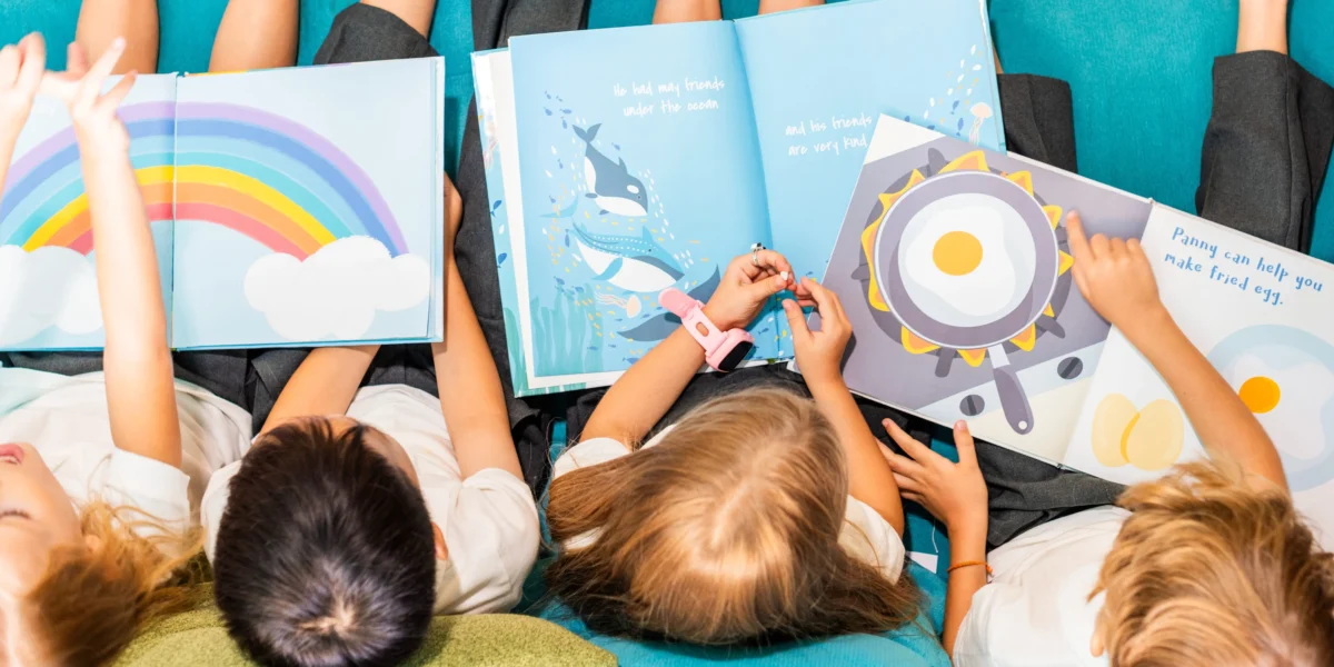 Top-down view of children lying on a teal carpet, reading bright picture books together with rainbow and ocean-themed illustrations.
