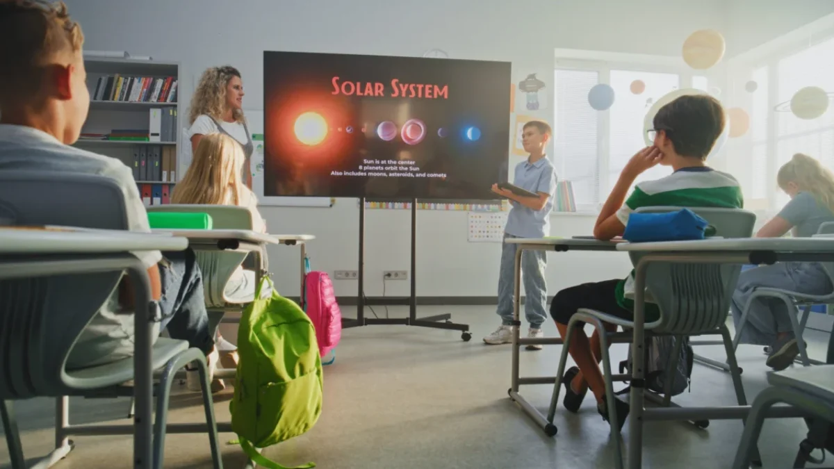Teacher leads a solar system lesson as students sit at desks facing a large screen in a bright classroom.