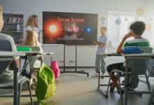 Teacher leads a solar system lesson as students sit at desks facing a large screen in a bright classroom.
