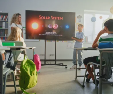 Teacher leads a solar system lesson as students sit at desks facing a large screen in a bright classroom.
