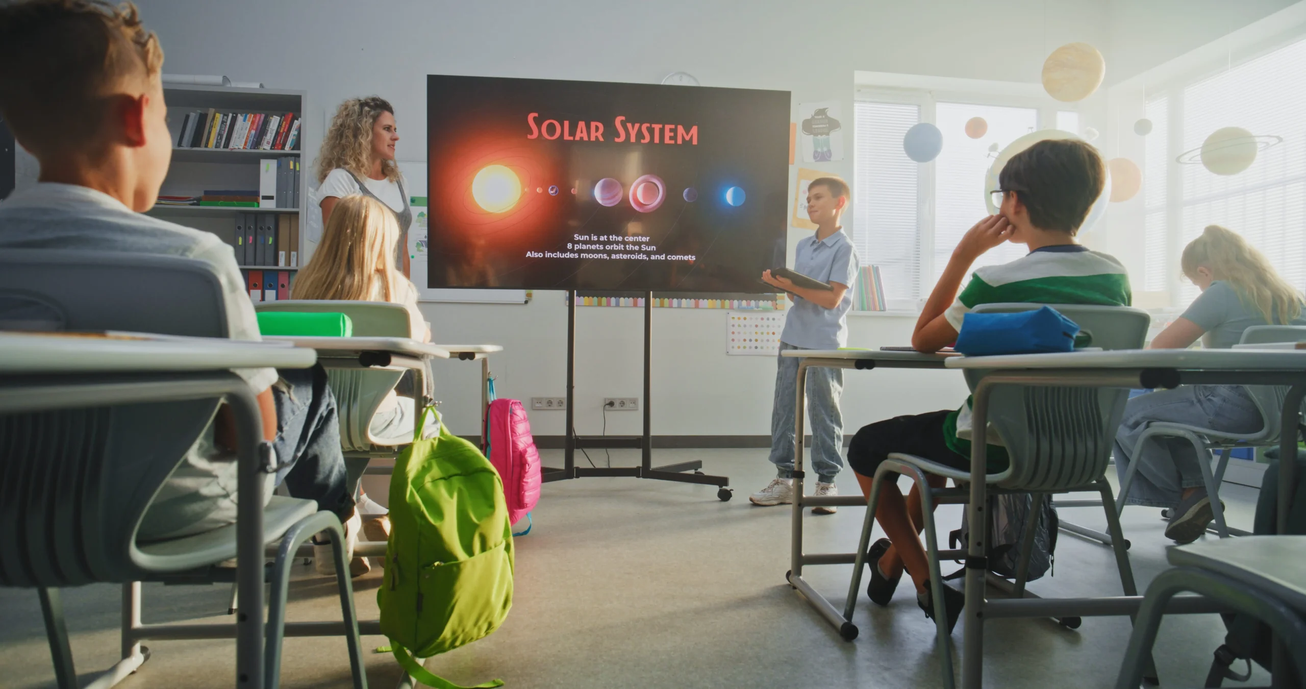 Teacher leads a solar system lesson as students sit at desks facing a large screen in a bright classroom.