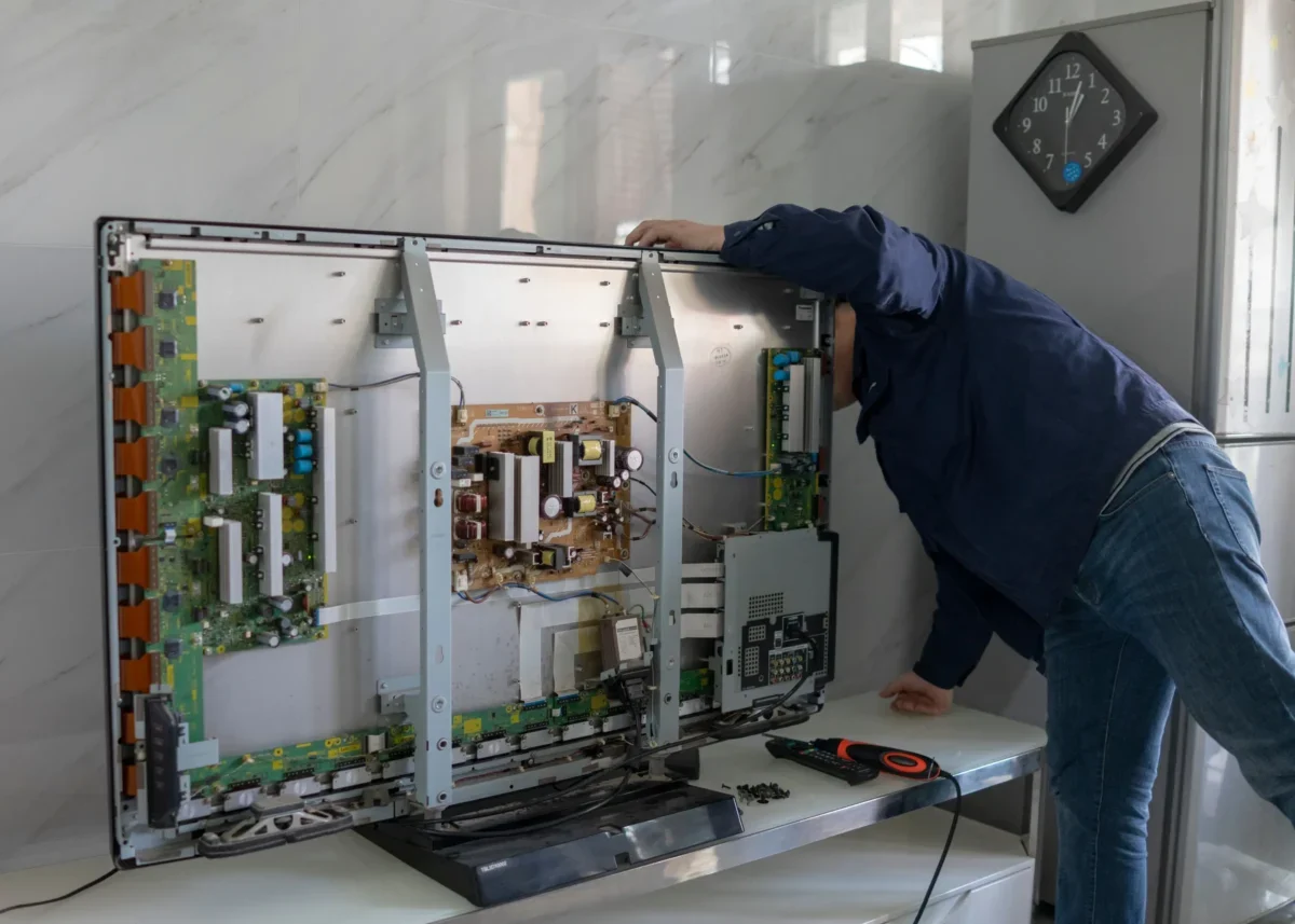Technik v modré košili opravuje odkrytou elektronickou skříň s deskami plošných spojů uvnitř skříně; v pozadí viditelné nástěnné hodiny