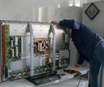 Technik v modré košili opravuje odkrytou elektronickou skříň s deskami plošných spojů uvnitř skříně; v pozadí viditelné nástěnné hodiny