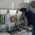 Technik v modré košili opravuje odkrytou elektronickou skříň s deskami plošných spojů uvnitř skříně; v pozadí viditelné nástěnné hodiny