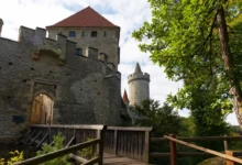 Kamenná hradní zeď s kulatou věží a červenou taškovou střechou, pohled z dřevěné lávky mezi zelenými stromy.