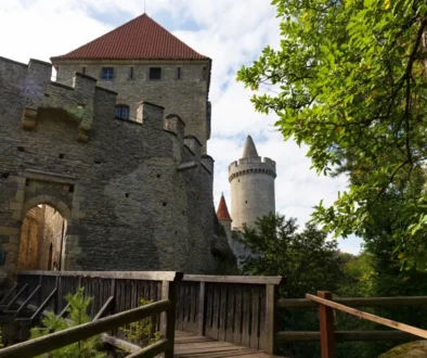 Kamenná hradní zeď s kulatou věží a červenou taškovou střechou, pohled z dřevěné lávky mezi zelenými stromy.